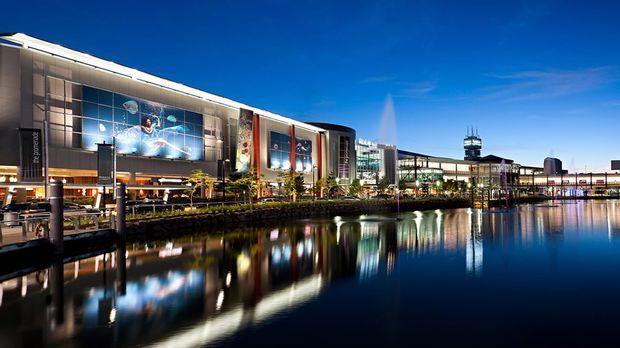 Nighttime waterfront view of a large contemporary commercial and entertainment complex, with illuminated glazed facades, prominent graphic displays, landscaped pedestrian edges, and a calm reflecting body of water in the foreground.