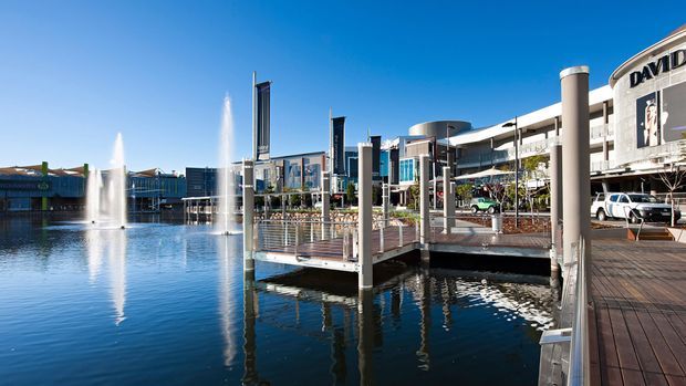A bright daytime view of a waterfront commercial precinct with a broad ornamental pond, fountains, pedestrian boardwalks, and contemporary multi-storey retail or mixed-use buildings. The scene features extensive glazing, sculptural vertical elements, landscaped public spaces, and clear blue skies.