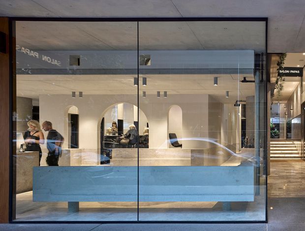 A contemporary commercial salon interior viewed through a large full-height glazed storefront. The space features pale stone or plaster finishes, arched wall openings, minimalist black track and pendant lighting, and a substantial light-blue reception counter in the foreground. Salon stations and people are visible inside, with a neighbouring illuminated stairway and signage at the right.