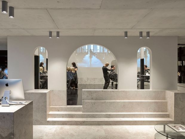 A refined commercial interior with a pale stone reception counter and stepped central platform. The space features arched wall openings, extensive light marble or stone surfaces, exposed concrete ceilings, recessed cylindrical spotlights, dark shelving, and a projected image on the rear wall. People and workstations are visible through the openings and reflections, suggesting a design studio, showroom, or gallery-like workplace.