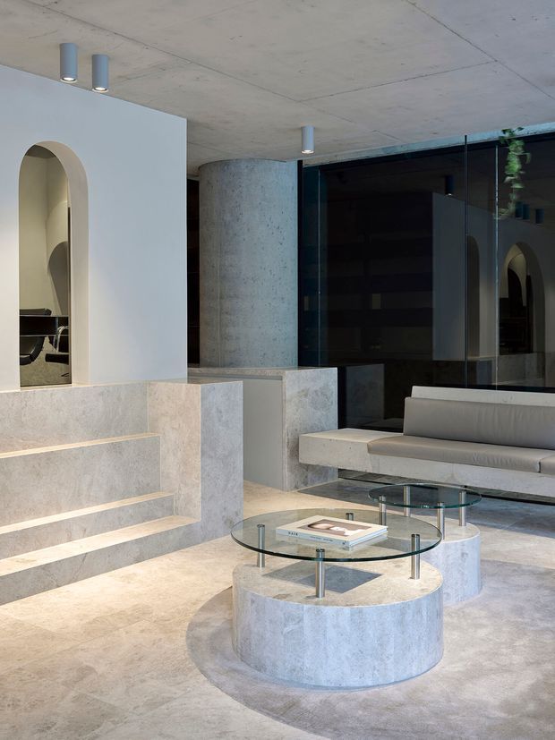 A refined contemporary interior lounge or reception space with a pale concrete and stone palette. Built-in stepped seating rises along the left wall beside a rounded arch opening, while a cylindrical concrete column and dark full-height glazed partition define the rear. A sculptural light upholstered sofa faces two round glass-topped, cylindrical concrete coffee tables. Exposed concrete ceilings, recessed and suspended cylindrical lighting, and minimal furnishings create a polished yet monolithic atmosphere.