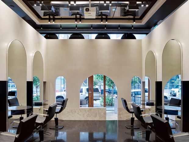A symmetrical, double-height commercial salon interior with black styling chairs and workstations arranged along both sides. Tall arched mirrors and arched openings define the pale plaster walls, while a polished dark stone or terrazzo floor reflects the space. The ceiling features exposed dark structural framing, track lighting, and a large mirrored or reflective inset. Full-height glazing and a central entrance overlook the street outside.