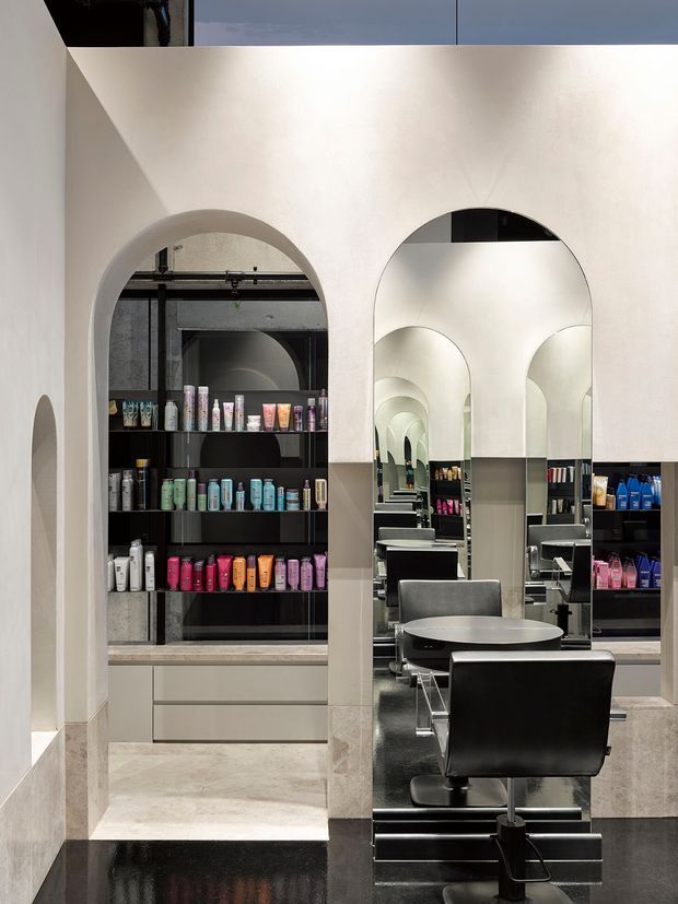 Interior view of a contemporary hair salon with a refined monochromatic fit-out. Cream plaster walls feature large rounded arch openings and an arched full-height mirror, creating repeated reflections through the salon. Black styling chairs, counters, shelving, and glossy dark flooring contrast with pale stone-look surfaces and neatly arranged retail products.