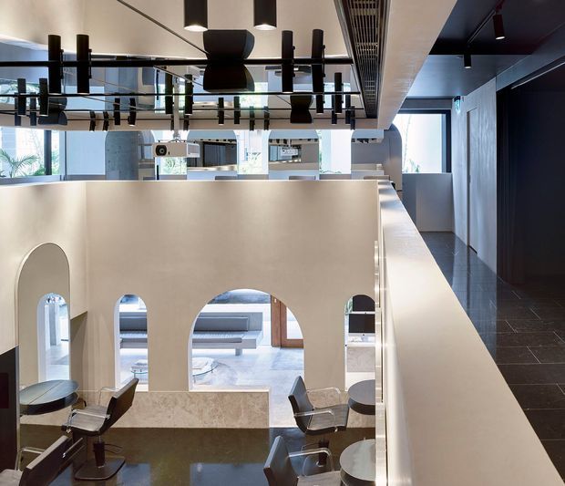A contemporary multi-level commercial interior, likely a salon or hospitality space, viewed from an upper walkway overlooking a lower seating and service area. The space features large arched wall openings, pale plaster walls, dark tiled flooring, stone counters, salon-style chairs, exposed services, and dramatic black pendant and track lighting. Reflective ceiling elements and abundant glazing create a bright, spacious atmosphere with strong geometric contrast.
