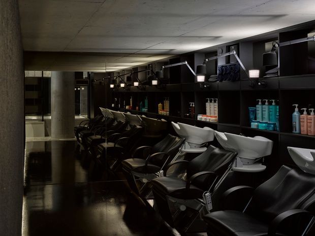 A dark, contemporary commercial salon interior with a long row of black shampoo stations and white ceramic wash basins. Extensive black built-in shelving above the stations displays salon products, with adjustable task lighting integrated into the joinery. The space has a low exposed concrete ceiling, polished dark flooring, and reflective surfaces, creating a moody, industrial atmosphere.