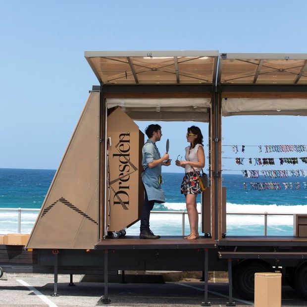 A branded mobile retail or display trailer parked beside a beach, with its side opened into a compact showroom. Two people stand inside among hanging display items, while the trailer has raised roof panels, fabric awnings, dark structural framing and support legs. Ocean, surf and a railing are visible in the background.
