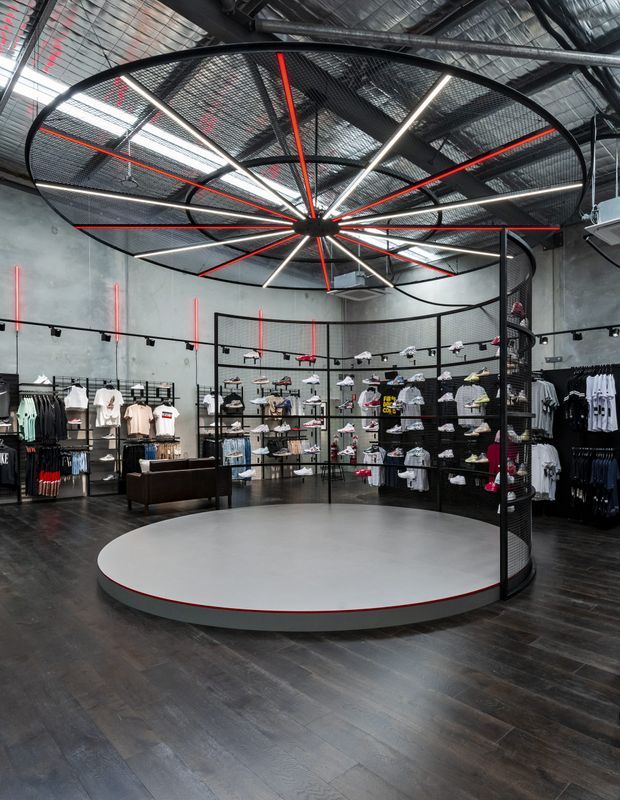 Interior view of a contemporary sportswear retail store with dark timber-look flooring, exposed industrial ceiling services, concrete walls, black mesh display structures, clothing racks, and a large circular raised platform at the centre. Dramatic black, white, and red linear lighting forms a radial feature overhead.