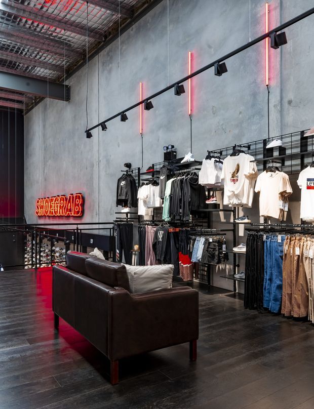 Interior of an urban fashion retail store with exposed industrial ceiling structure, raw grey concrete walls, dark timber flooring, black metal track lighting, wall-mounted clothing racks, and a dark leather lounge chair. Red neon tube lighting and signage provide vivid accents against the monochrome industrial fit-out.