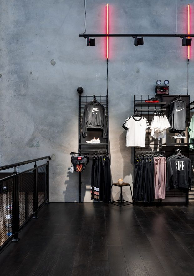 Interior of a contemporary sportswear retail store with clothing displayed on black metal grid wall systems. The space features a raw grey concrete wall, dark flooring, black metal railings and fixtures, and vertical red neon accent lighting.