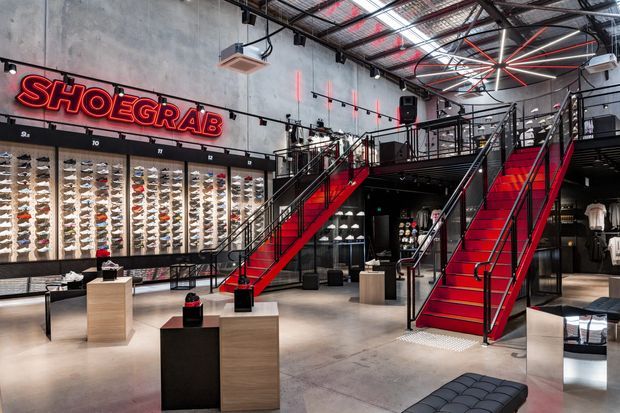 Interior view of a contemporary shoe retail store with an industrial-style fit-out. The large open showroom features exposed concrete walls, an exposed services ceiling, extensive illuminated shoe displays, black metal mezzanine structures, bold red staircases and railings, and light timber product plinths.