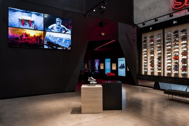 A dark, dramatic retail sneaker store interior with black walls, polished concrete flooring, illuminated wall-mounted shoe displays, digital screens, display plinths, and red accent lighting. The space has an industrial contemporary fit-out with a strong branded, gallery-like presentation.