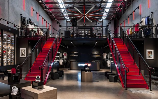 A dramatic two-level retail showroom with a central reception counter, symmetrical red staircases, black metal balustrades, exposed industrial ceiling services, concrete-look walls and floor, and clothing and footwear displays arranged along both sides. Bold linear and circular lighting features create a high-contrast, theatrical atmosphere.