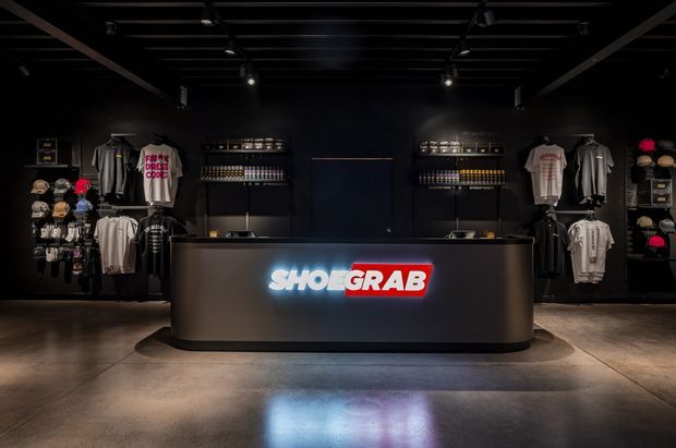 A dark, industrial-style retail showroom interior with a central curved reception or checkout counter featuring an illuminated SHOE GRAB logo. Black shelving and wall displays hold apparel, shoes, caps, and accessories, while exposed dark ceiling beams, track lighting, and a polished concrete floor create a dramatic commercial fit-out.
