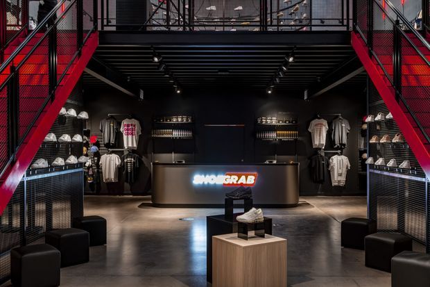 A dark, industrial-style retail interior with a centrally positioned illuminated reception or checkout counter, merchandise displays along both side walls, black seating, and a shoe display pedestal in the foreground. Red metal staircases and black grated mezzanine walkways frame the space, while exposed dark ceiling structure and track lighting reinforce the dramatic athletic-fashion store aesthetic.