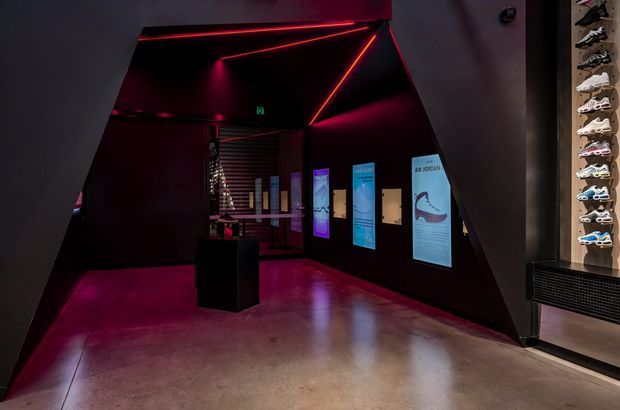 A dark, immersive commercial exhibition or retail interior with angular black architectural walls, polished concrete flooring, and vivid red linear LED lighting. Illuminated informational display panels line the walls, while a vertical sneaker display is visible at the right edge. The space has a dramatic, gallery-like atmosphere.