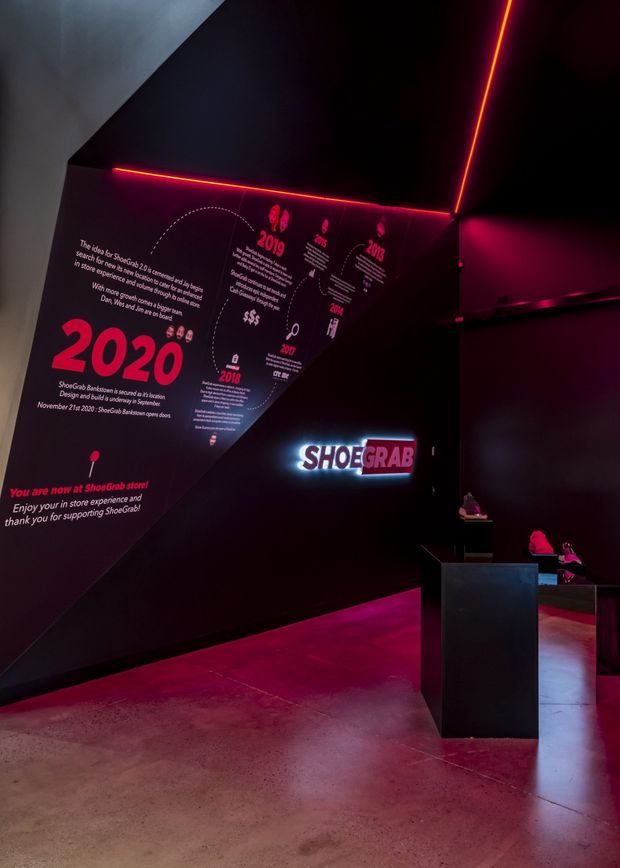 Dark, immersive retail interior featuring angular black walls, magenta and red linear LED lighting, polished concrete flooring, and illuminated Shoegrah branding. Large graphic wall signage presents a company timeline, while black display plinths showcase footwear and products in the background.