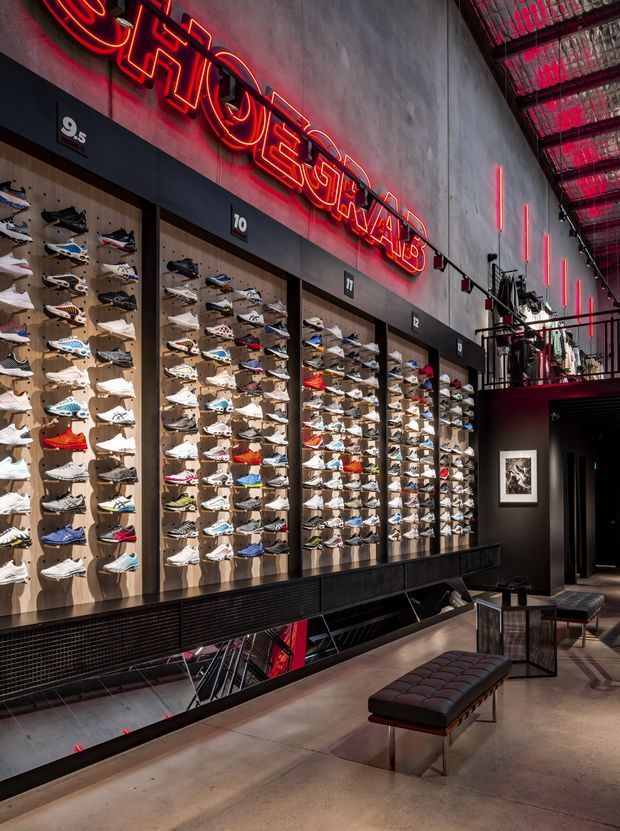 Interior of a contemporary sneaker retail store with extensive wall-mounted shoe displays, dark metal framing, concrete finishes, dramatic red neon signage and accent lighting, and a small lounge area with a tufted bench and side tables. The space has an industrial, high-contrast commercial design with an open view toward a lower level.