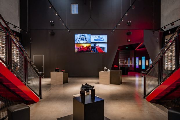A dramatic contemporary exhibition or gallery interior with a dark charcoal palette, polished concrete floor, red illuminated staircases on both sides, black metal mesh balustrades, track lighting, display plinths, and wall-mounted digital screens. The symmetrical space has an industrial, immersive character and appears to include an entrance or further gallery area at the rear.