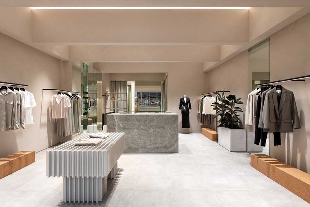 A refined contemporary clothing boutique with pale stone flooring, warm neutral walls, exposed ceiling beams, integrated linear lighting, mirrored display panels, and minimalist clothing racks. A central textured stone display island and a ribbed light-toned table anchor the spacious retail interior, with tailored garments arranged along both sides and a potted plant adding greenery.