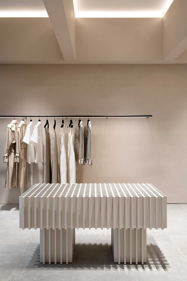 A minimalist retail or fashion display space with a neutral beige interior, exposed ceiling beams and concealed linear lighting. A clothing rail displays neutral-toned garments behind a sculptural slatted display table or island, with a pale tiled floor and strong geometric shadows.