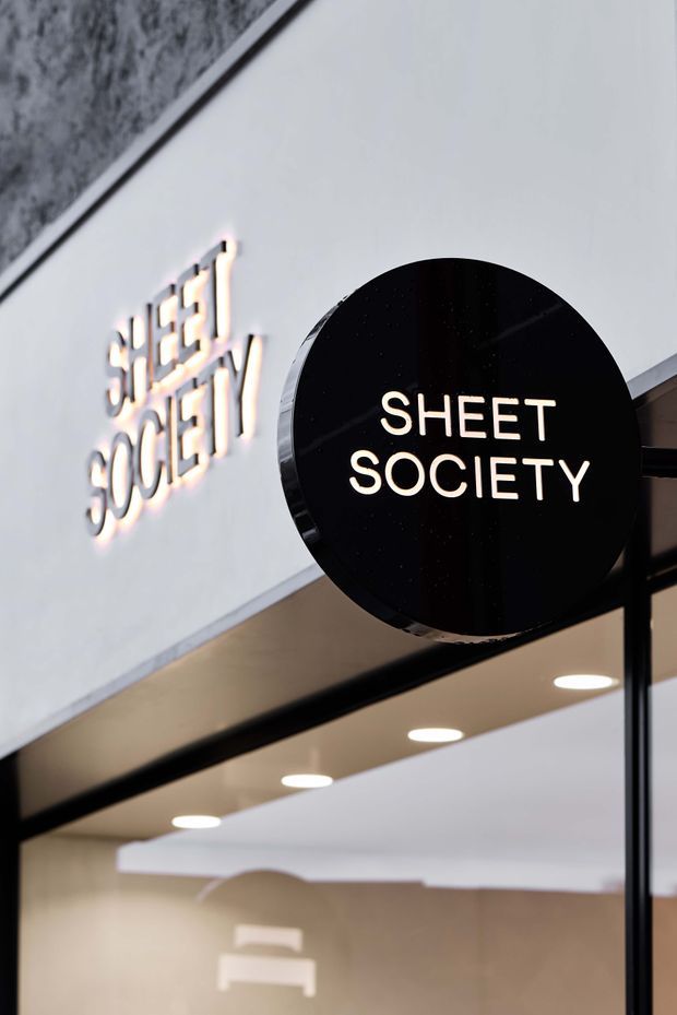 Close-up architectural detail of a contemporary retail storefront featuring a black circular projecting sign reading “SHEET SOCIETY,” illuminated lettering, black-framed glazing, recessed ceiling downlights, and a neutral-toned interior visible beyond the entrance.