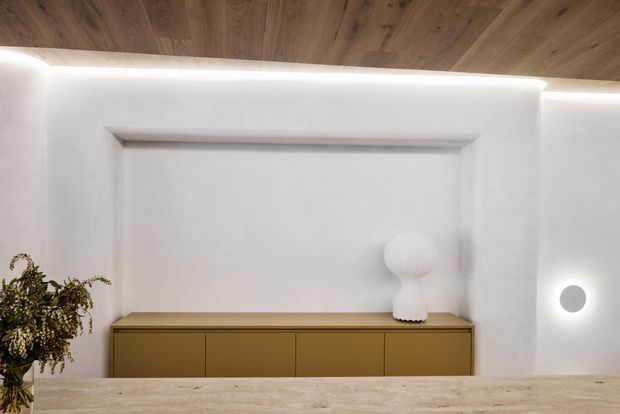A refined minimalist interior vignette featuring a long mustard-brown built-in cabinet beneath a recessed white wall niche. Warm timber ceiling boards, softly illuminated concealed perimeter lighting, a sculptural white vase, and a small plant create a calm, contemporary atmosphere. A pale stone or timber-toned surface occupies the foreground.
