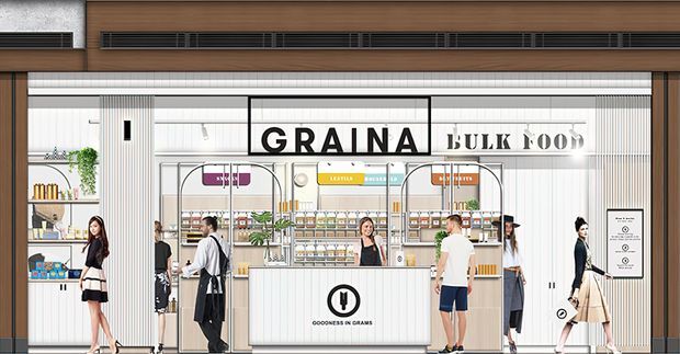 Street-facing retail storefront for a grain-focused bulk food shop, with a prominent GRAINA BULK FOOD sign, bright white tiled and ribbed wall finishes, timber-toned upper facade, glass frontage, and a central pale wood retail counter. Shelving displays packaged goods and bulk dispensers inside, with several shoppers visible near the entrance.