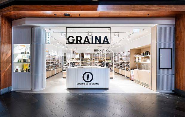 Front-facing view into a bright retail food shop branded “GRAINA,” with a central white checkout or service counter, extensive shelving of packaged goods, white ribbed cylindrical columns, track lighting, and a dark tiled floor. The shop entrance is framed by substantial warm-toned timber beams and dark trim, creating a contrast between the bright minimalist interior and the darker threshold.