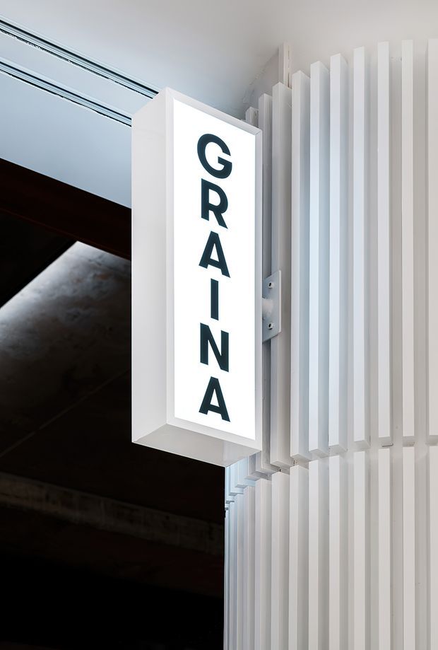 Close-up architectural detail of a commercial facade or entrance featuring a vertically illuminated sign reading “GRAIN,” mounted beside a white three-dimensional slatted screen. Dark structural elements and a linear ceiling detail are visible in the background.
