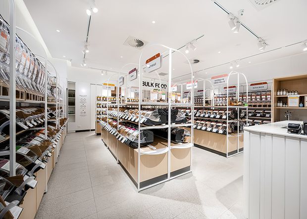 Bright modern bulk-food retail store interior with extensive wall shelving and central freestanding dispensing units. The space has a white ceiling with recessed and track lighting, light wood-toned cabinetry, pale grey tiled flooring, white counters, and clear category signage throughout.