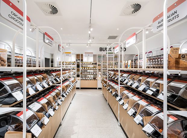 Bright, modern bulk-food retail interior viewed down a central aisle. Rows of wooden-based dispensing and display bins hold packaged dry goods, with white metal shelving frames, curved overhead category signs, a white ceiling with recessed spotlights and ventilation grilles, and a light terrazzo-style floor. The space has a clean, highly organised supermarket or bulk-goods store layout.