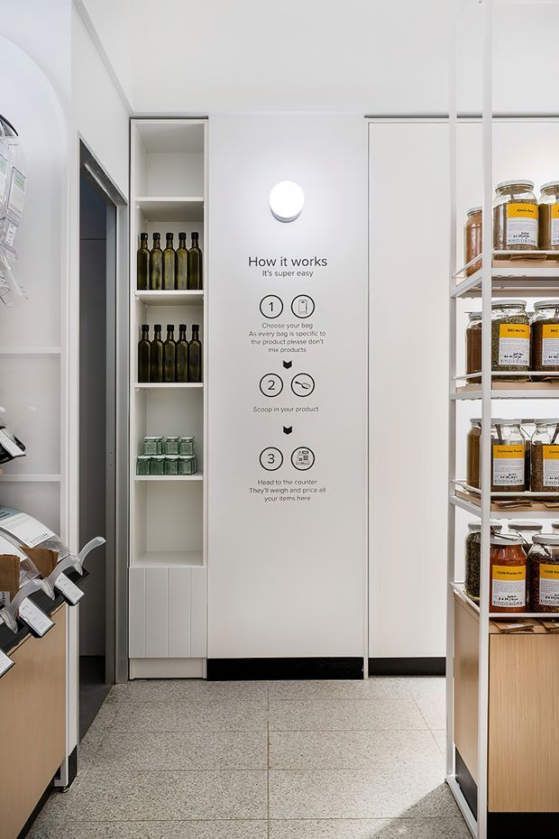 Bright, minimalist commercial refill store or pantry-like retail interior, viewed down a narrow aisle. White floor-to-ceiling cabinetry and shelving frame jars of dry goods and bottles, with a central white panel displaying illustrated instructions for selecting and scooping products. The space has pale terrazzo-style flooring, integrated lighting, and light timber cabinetry at the lower shelving units.