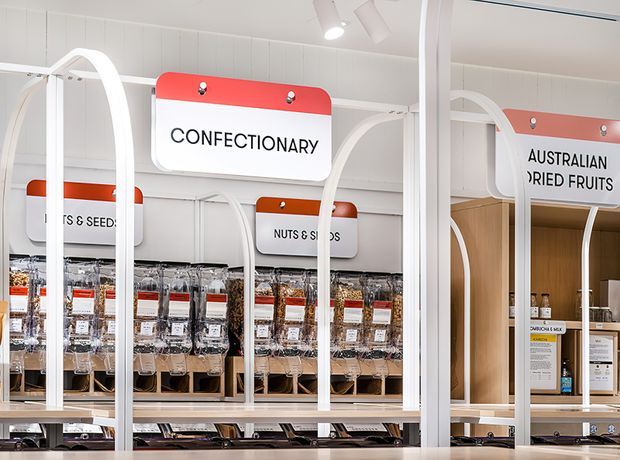 Bright, contemporary bulk-food retail interior with white arched display frames, overhead category signage, transparent dispensers filled with nuts and dried goods, and clean integrated shelving and counters. The space has a crisp white palette with red and orange signage accents and strong linear lighting.