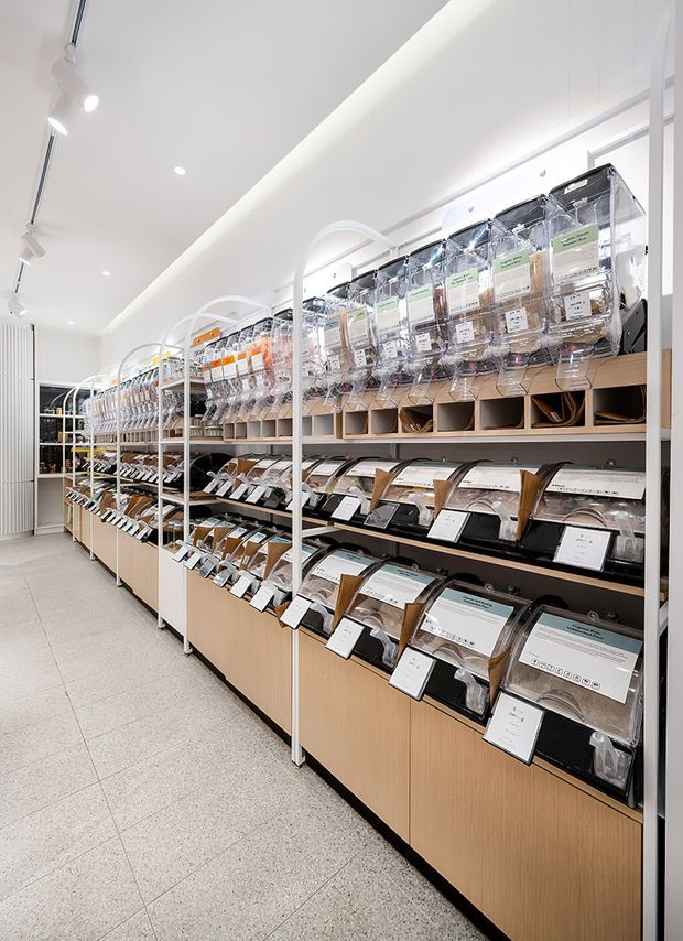 Bright, modern retail showroom interior with long rows of organised sample displays in clear containers and angled presentation trays. The space has white walls and ceiling, linear track lighting, pale timber-toned built-in cabinetry, black shelving, and a light terrazzo- or tile-like floor. The image is dominated by product/sample merchandising and fixed display joinery.
