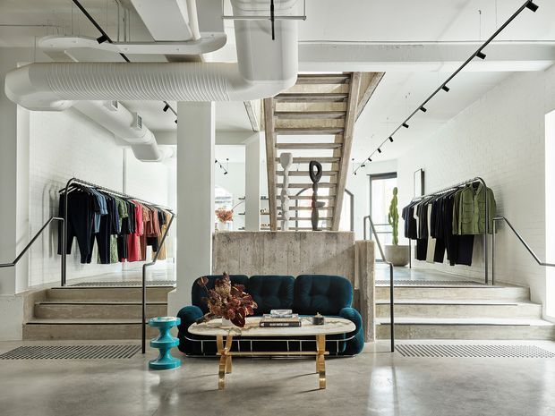 Bright, open-plan fashion retail interior in a converted industrial-style space. Clothing racks line both sides of a central lounge area with a dark teal curved sofa, oval coffee table, decorative objects, and a turquoise side table. A raw concrete half-wall and exposed stair structure lead to an upper level, while white-painted brick walls, exposed white ductwork, polished concrete floors, and black track lighting reinforce the contemporary industrial character.