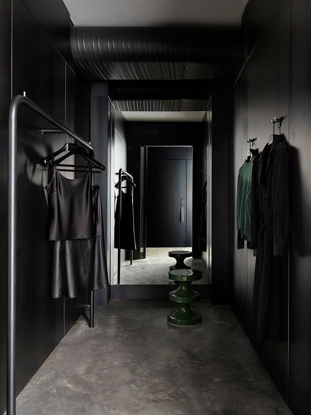 A narrow, dark dressing corridor or wardrobe area with black wall panels, exposed black ductwork, wall-mounted clothing rails and hooks, and a full-height mirror at the far end. The space has a polished industrial character, with a grey concrete-look floor and sculptural green and black stools placed centrally.