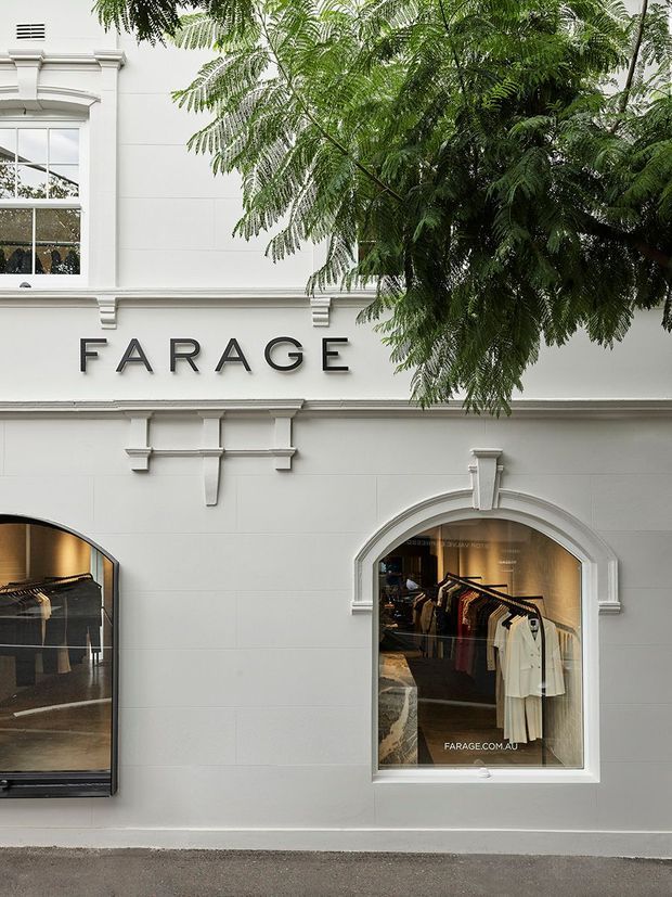 Street-facing facade of a refined fashion retail storefront branded “FARAGE.” The white rendered building features classical architectural detailing, moulded cornices, pilasters, decorative window surrounds, and arched display windows. Clothing racks and garments are visible through the glazing, with a leafy tree partially obscuring the upper facade.