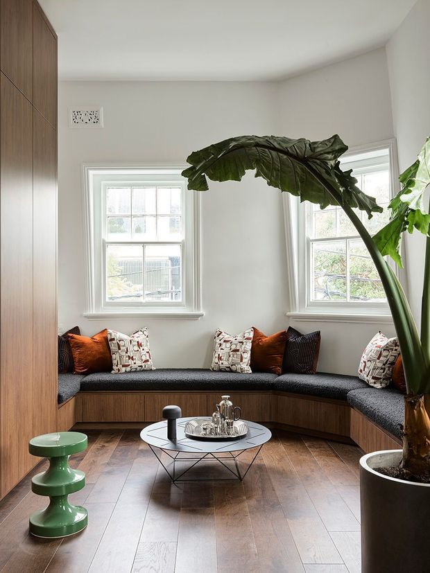 Bright, compact sitting room with a curved built-in window seat upholstered in charcoal fabric, integrated beneath two sash windows. Warm timber cabinetry and paneling line the left and lower walls, complemented by patterned and rust-coloured cushions. A round wire-base coffee table, green sculptural side table, and large potted tropical plant add contemporary character. The room has white walls and ceiling, natural timber flooring, and abundant daylight.