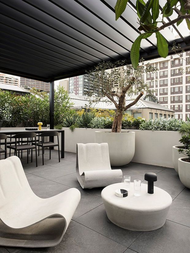 A refined rooftop terrace with a dark slatted pergola, outdoor dining setting, sculptural white lounge chairs, a rounded coffee table, and large planters filled with mature greenery. The terrace overlooks a dense urban setting and uses a restrained palette of charcoal grey, white, and green with strong contemporary lines.