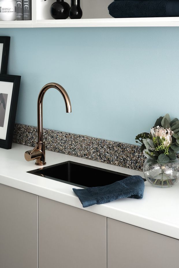 Close-up of a contemporary kitchen or utility-room sink area with a matte white benchtop, integrated black undermount sink, bronze curved mixer tap, taupe-grey handleless cabinetry, pale blue wall, and a narrow terrazzo-look splashback. Dark blue towels and decorative shelving add contrast.