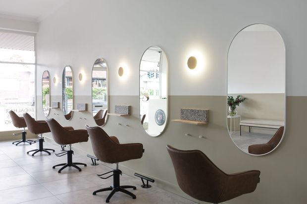 A bright, refined hair salon interior with a row of brown swivel styling chairs facing large arched mirrors. The walls are painted in a two-tone scheme with a light upper section and muted taupe lower section, complemented by warm glowing wall sconces and small upholstered wall-mounted shelves. Large windows provide natural light, and the pale tiled floor contributes to the clean, airy atmosphere.