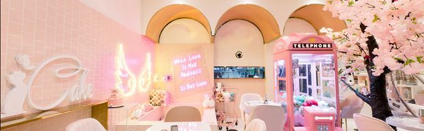 A highly stylised pink salon or hospitality interior with glossy pink tiled walls, arched ceiling niches, neon signage, pink-and-white furnishings, a vintage-style telephone booth, and artificial cherry-blossom decor. The space has a playful, bright, maximalist aesthetic with strong pastel pink and peach tones.