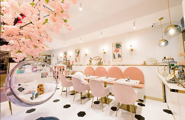 Bright, highly styled dessert café or patisserie interior with a predominantly white and blush-pink palette. The space features a long banquette with scalloped pink upholstery, pink dining chairs, white tables, patterned monochrome floor tiles, framed botanical artwork, decorative shelving, pendant lighting, and a prominent hanging bubble chair. A display counter and cake-service area are visible on the right, with floral decorative branches overhead.