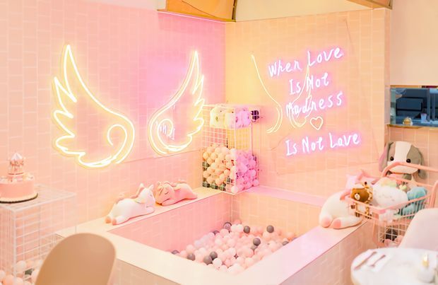 A whimsical pastel pink tiled bathroom with a sunken ball-pit-style bath or play tub filled with pastel balls. The walls feature glowing wing-shaped neon artwork and a handwritten neon quote, while plush toys, soft pastel accessories, and a small cake-like decorative object create a playful, highly stylised setting. The space has warm pink and peach lighting and a compact, immersive design.