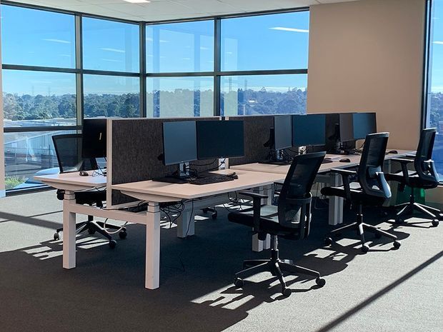 Bright open-plan commercial office with rows of white workstations, dark acoustic desk dividers, multiple computer monitors, and black ergonomic task chairs. Floor-to-ceiling corner glazing provides extensive views and strong natural light. The space has a low suspended ceiling, carpeted floor, and a clean, functional contemporary workplace fit-out.
