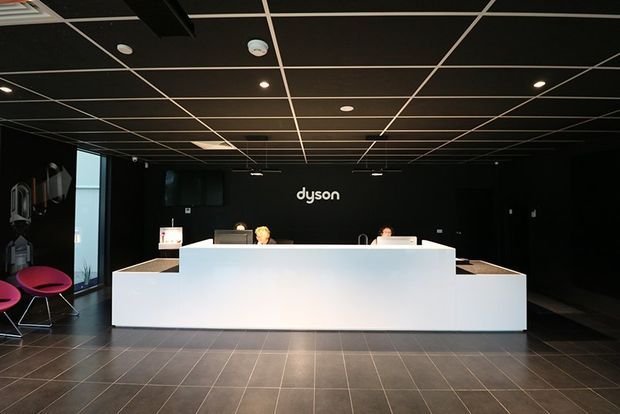 A dark, minimalist commercial reception area with a large angular white reception desk centered against a black wall bearing the Dyson logo. The space has a black exposed grid ceiling with recessed lighting, dark tiled flooring, black walls, and a small seating area with bright pink chairs at the left. Reception staff and computer equipment are positioned behind the desk.