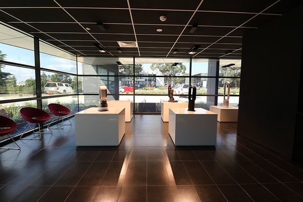 A contemporary commercial showroom or gallery interior with a dark tiled floor, black suspended grid ceiling, extensive floor-to-ceiling glazing, and white freestanding display plinths showcasing sculptural objects. Red lounge chairs line the glazed perimeter, with a bright street-facing exterior visible beyond the windows.