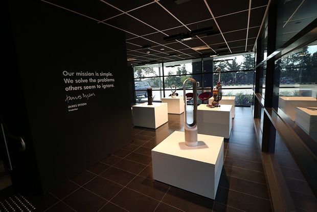 A dark, contemporary product showroom or commercial display space with a black feature wall bearing a mission statement, white pedestal plinths, and sculptural appliances or objects arranged for viewing. Floor-to-ceiling glazing lines the far and right sides, allowing views of trees and a roadway outside. The room has a black grid ceiling, dark tiled flooring, and a restrained black-and-white palette with warm daylight.