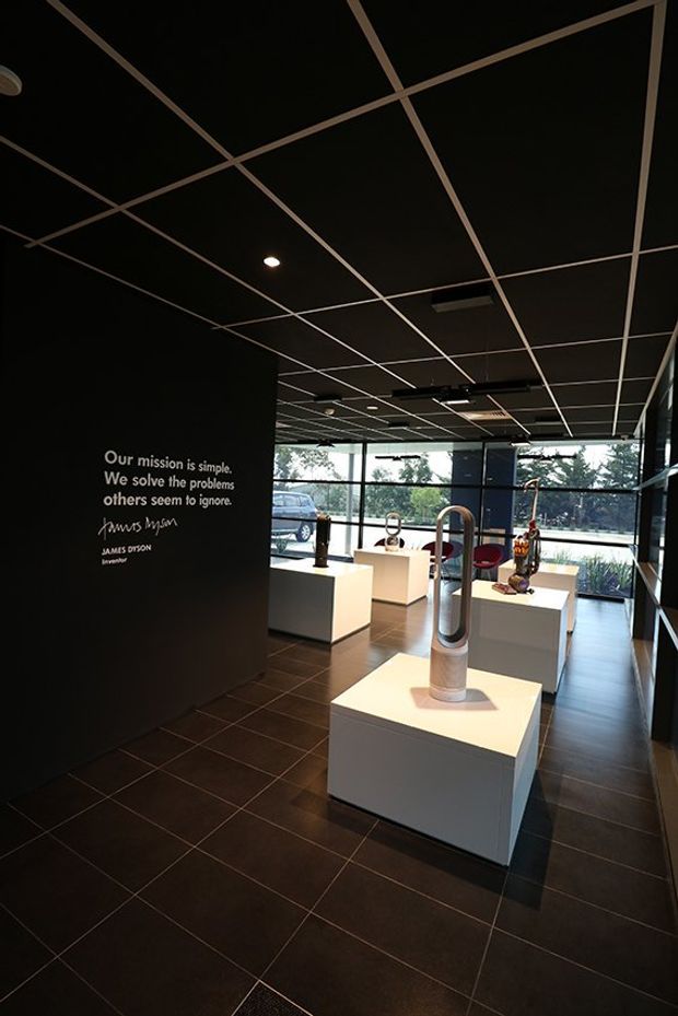 A dark, polished exhibition showroom with black walls and a black suspended grid ceiling, displaying sculptural vacuum cleaners and appliance products on white square plinths. Large floor-to-ceiling windows bring in daylight at the rear, while recessed and track lighting illuminates the displays. A James Dyson mission quote is printed on the feature wall, reinforcing the branded retail or product-gallery setting.