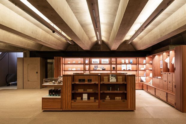 A warmly lit retail or display space with extensive custom timber shelving, cabinetry, glass-fronted display cases, and a central freestanding timber shelving unit. The ceiling features dramatic exposed concrete beams with linear recessed lighting and visible services. The overall interior is refined and gallery-like, with books, objects, and merchandise arranged throughout the built-in joinery.